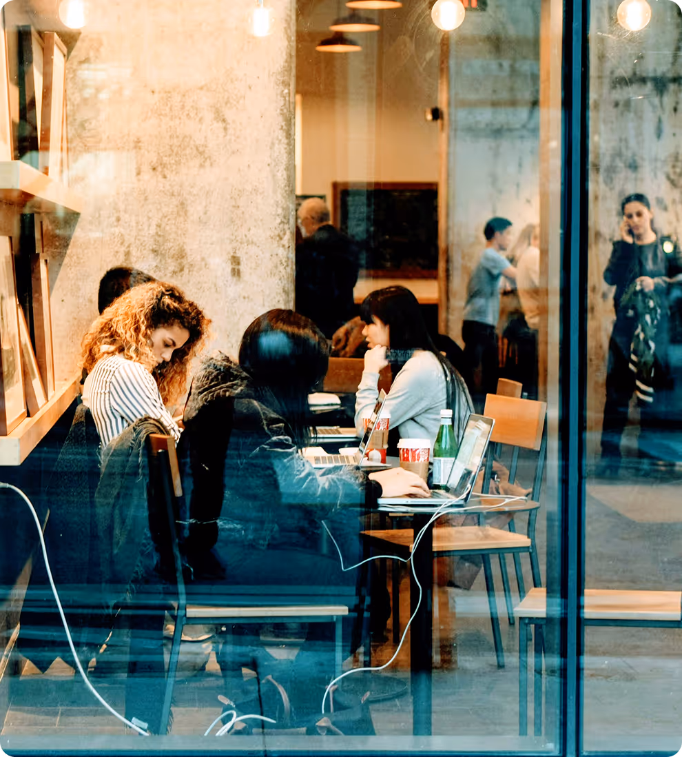 People working on laptops and drinking coffee inside a cafe viewed through a glass window.