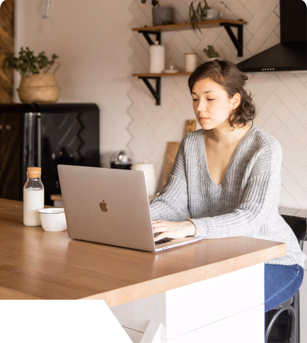 Woman in a gray sweater using a laptop at a wooden kitchen counter with a bottle and bowl nearby.
