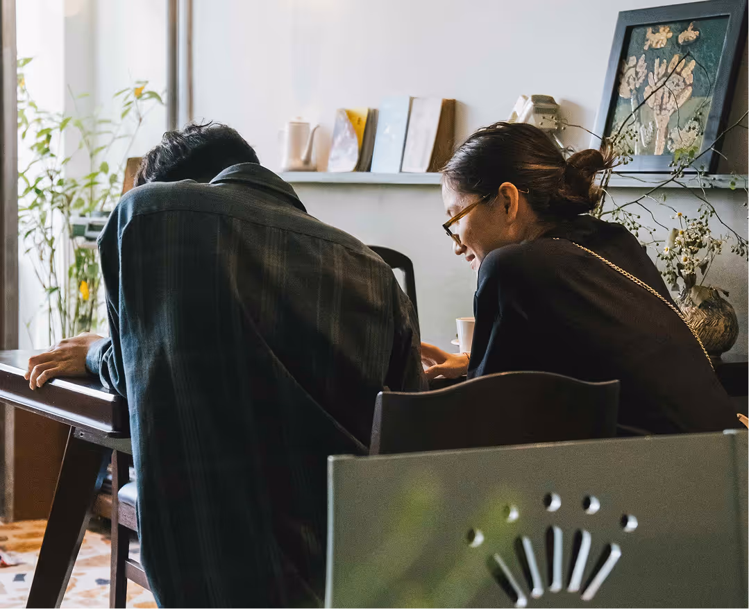 Two people sitting closely together at a desk, engaged in a conversation or collaborative activity in a cozy, plant-filled room.