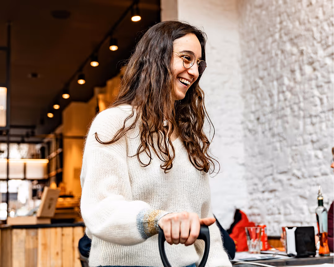 Smiling woman with long curly hair and glasses wearing a white sweater in a cozy indoor setting.