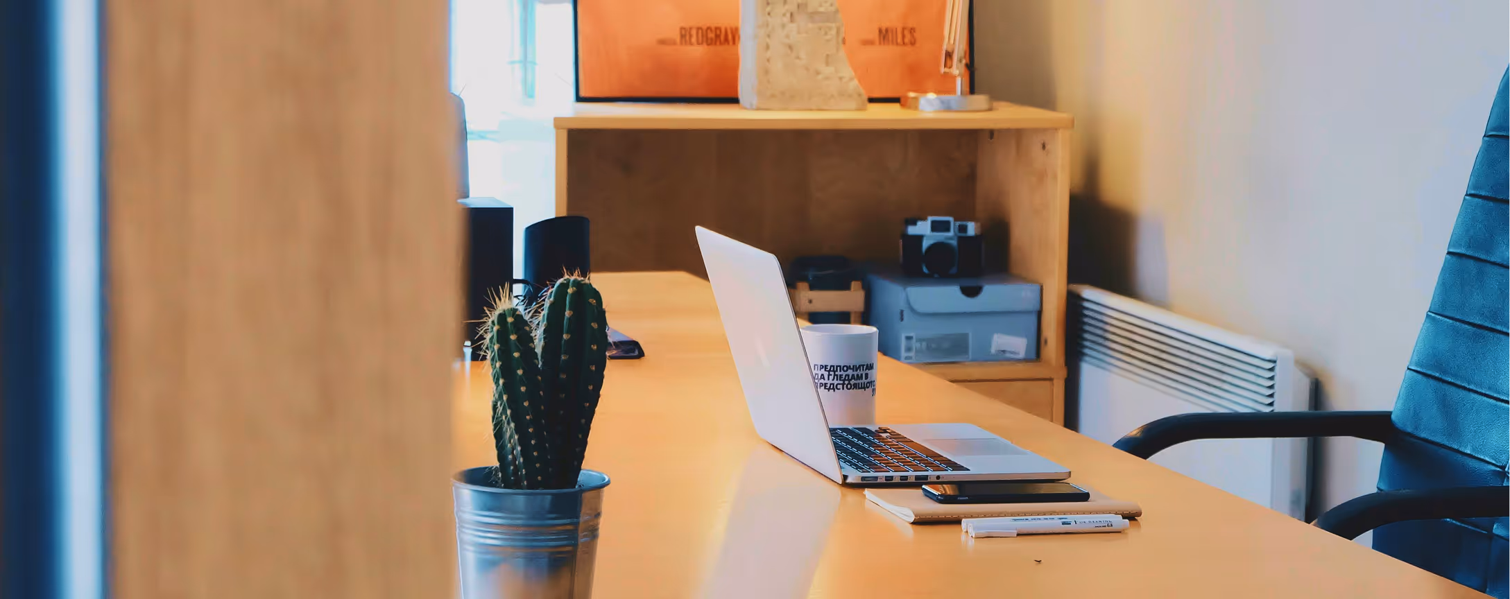 Workspace with a wooden desk featuring a potted cactus, an open laptop, a cup, a smartphone, pens, and a blue office chair.