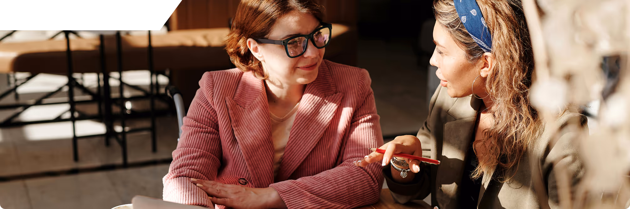 Two women engaged in a conversation at a table, one wearing glasses and a pink jacket, the other with a headband and holding a red pencil.