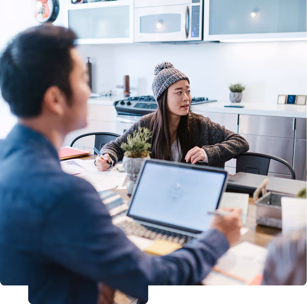 Two people having a discussion at a table with a laptop and papers in a modern kitchen setting.
