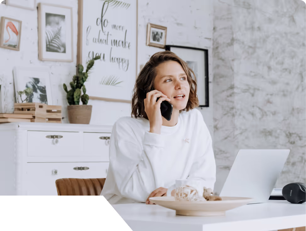 Woman in white sweater talking on phone while sitting at a white desk with laptop in a bright, decorated room.