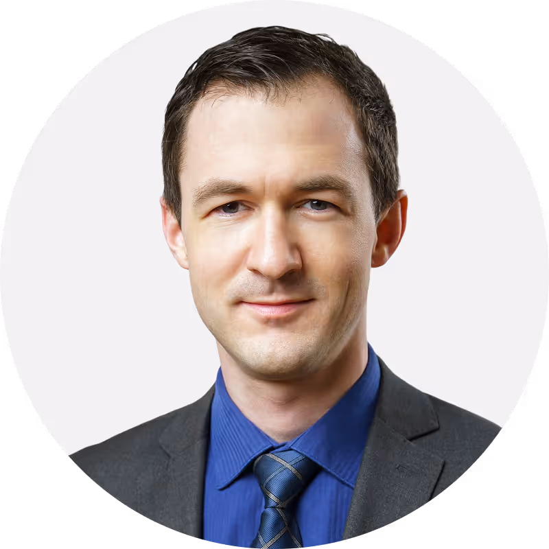 Portrait of a man wearing a dark suit, blue shirt, and striped tie, smiling slightly against a light background.