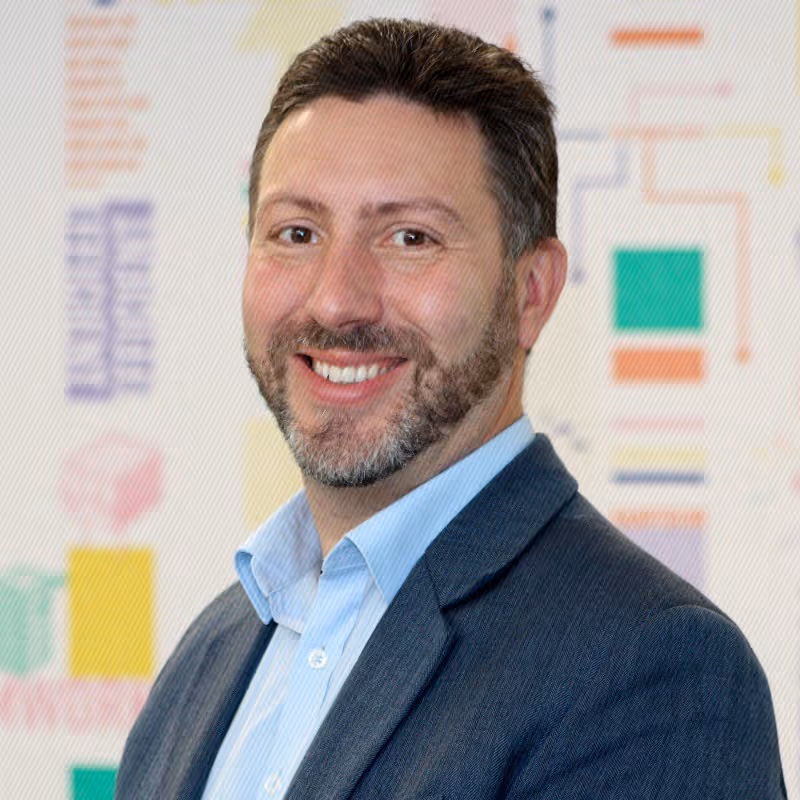 Smiling man with short dark hair and beard wearing a blue shirt and dark blazer, standing in front of a colorful blurred background.