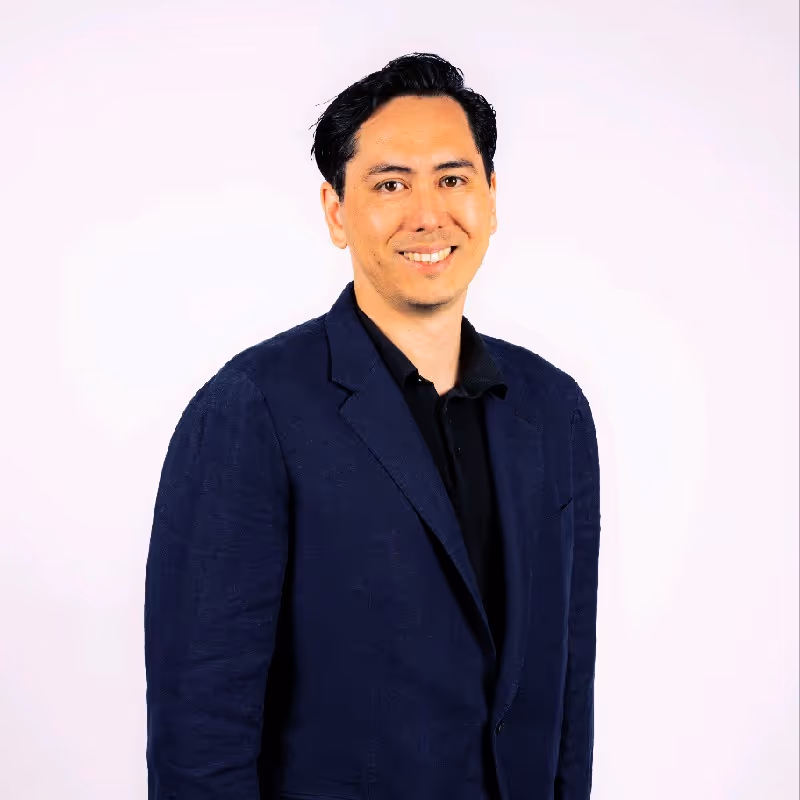 Smiling man with dark hair wearing a navy blazer and black shirt against a light background.