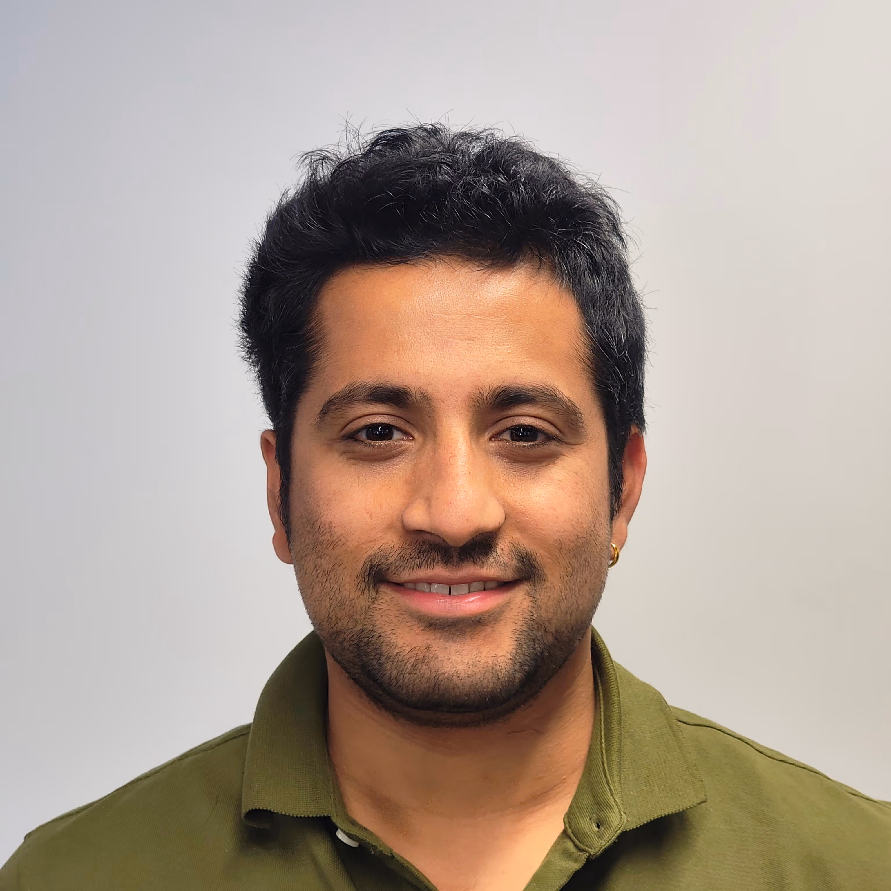 Smiling man with short curly black hair, trimmed beard, and gold hoop earring wearing an olive green collared shirt.
