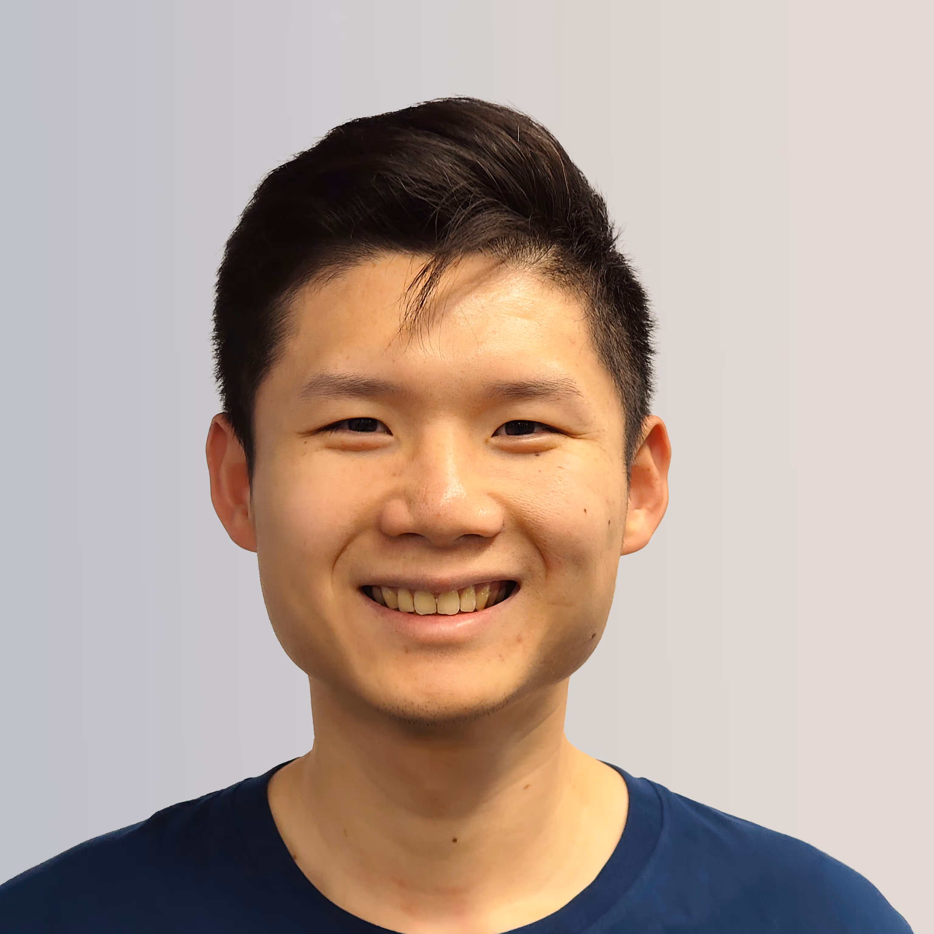 Young man with short black hair smiling wearing a dark blue shirt against a light gradient background.