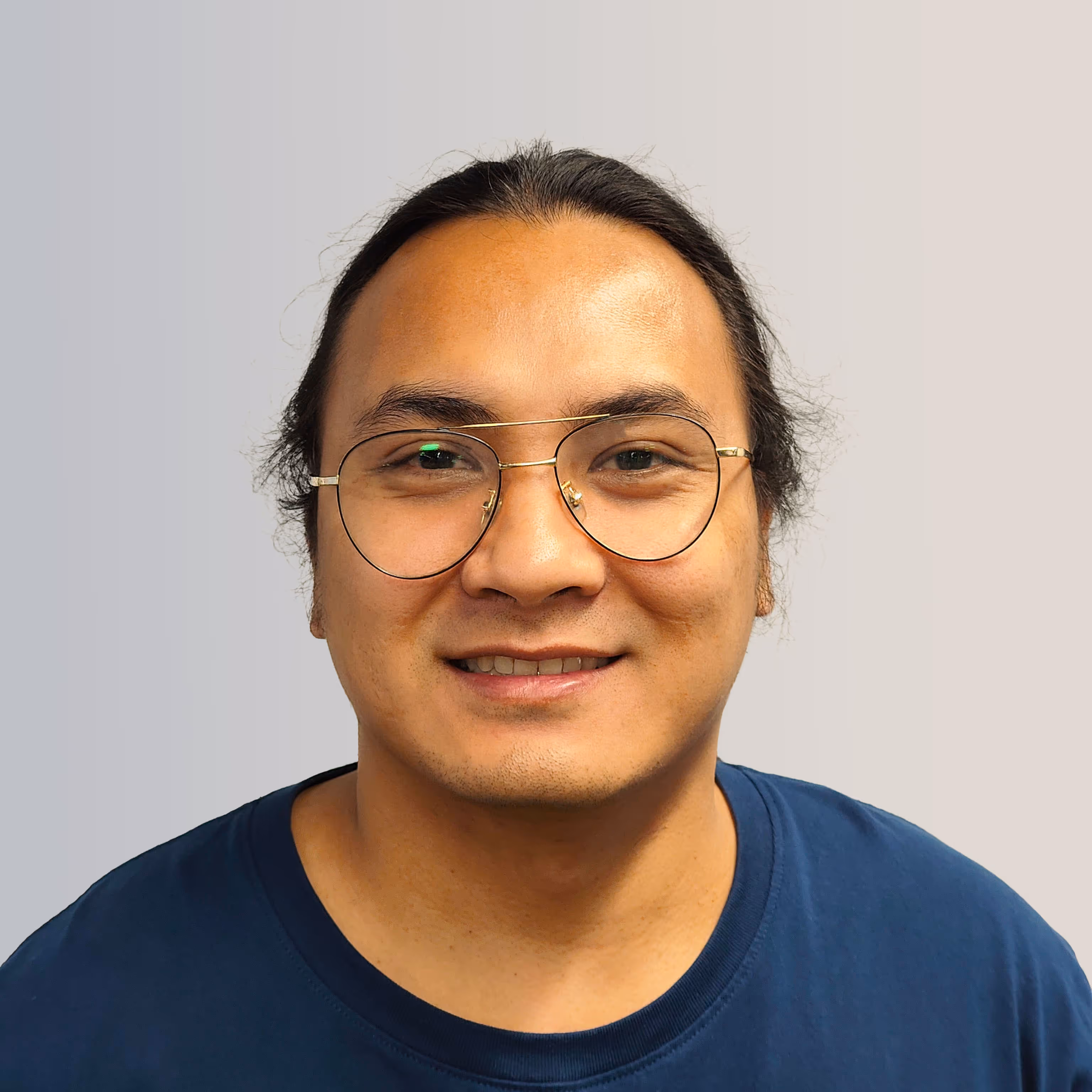 Smiling person with long black hair tied back wearing round glasses and a blue shirt against a light gray background.