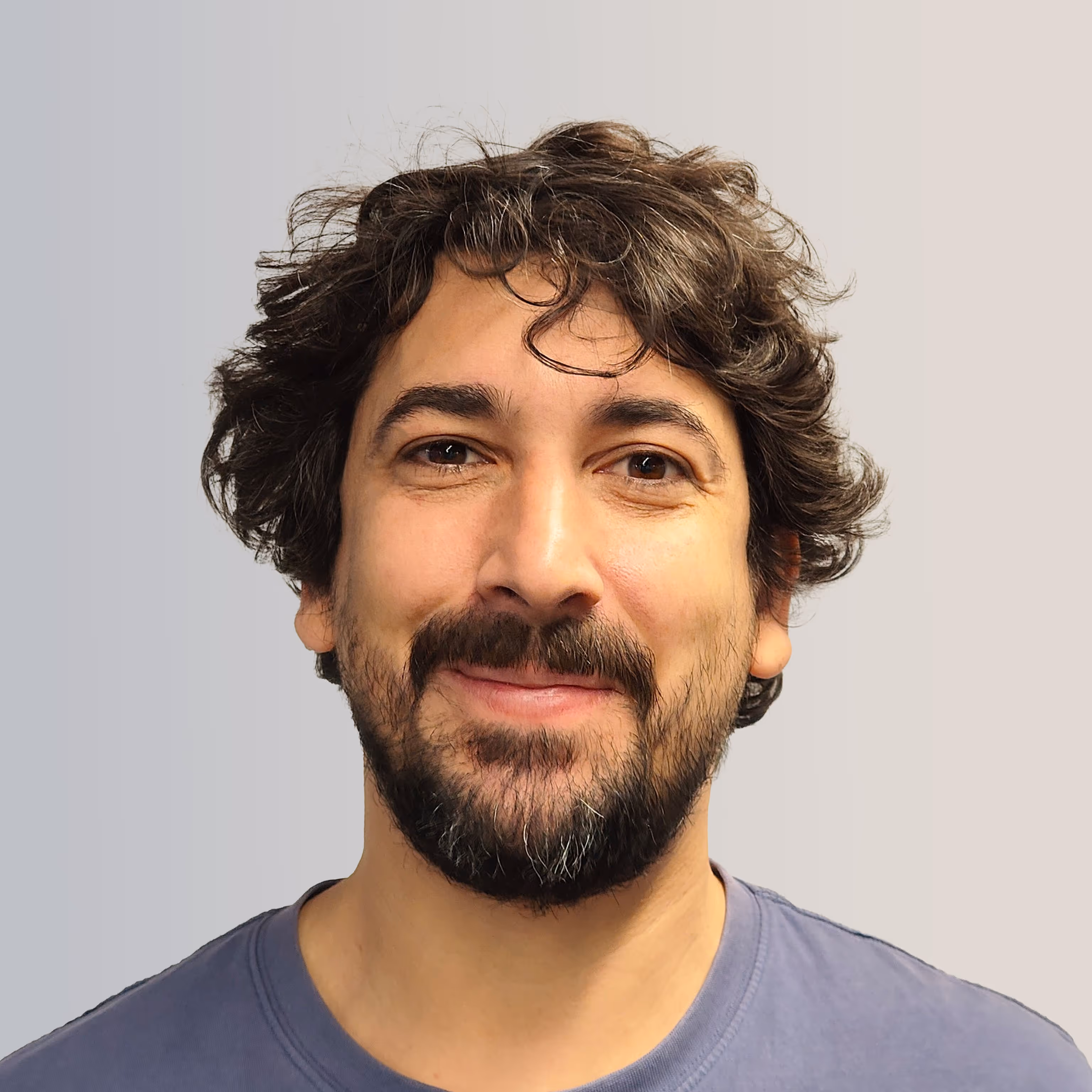 Man with curly dark hair and beard smiling against a light gray background.