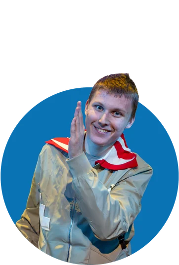 A smiling Tapestry actor in a tan jacket and red scarf gives a little wave