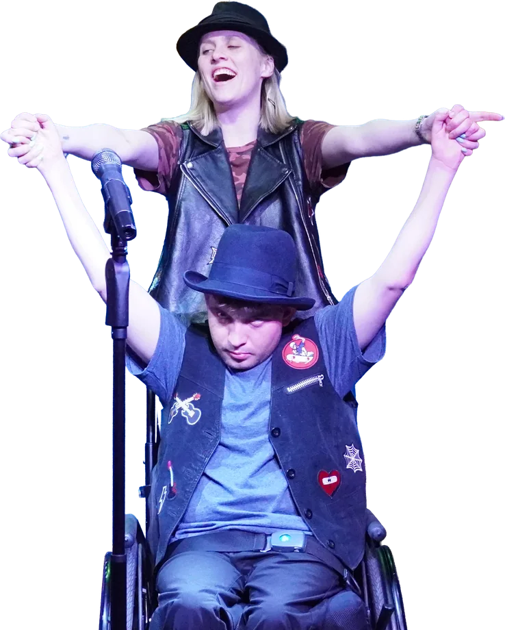 A Tapestry actor in a wheelchair raises his arms with his peer partner behind him, both in hats and vests