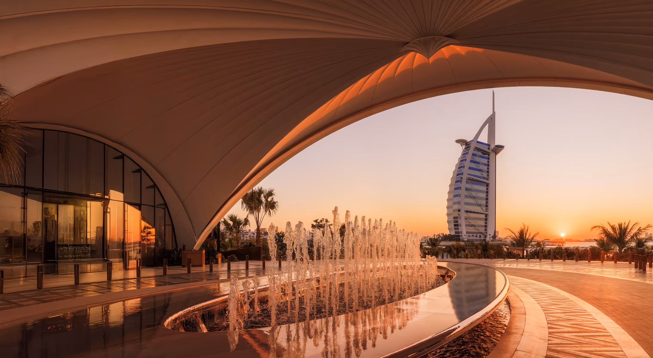 Sunset view of Burj Al Arab hotel framed by a curved architectural canopy and a fountain in Dubai.