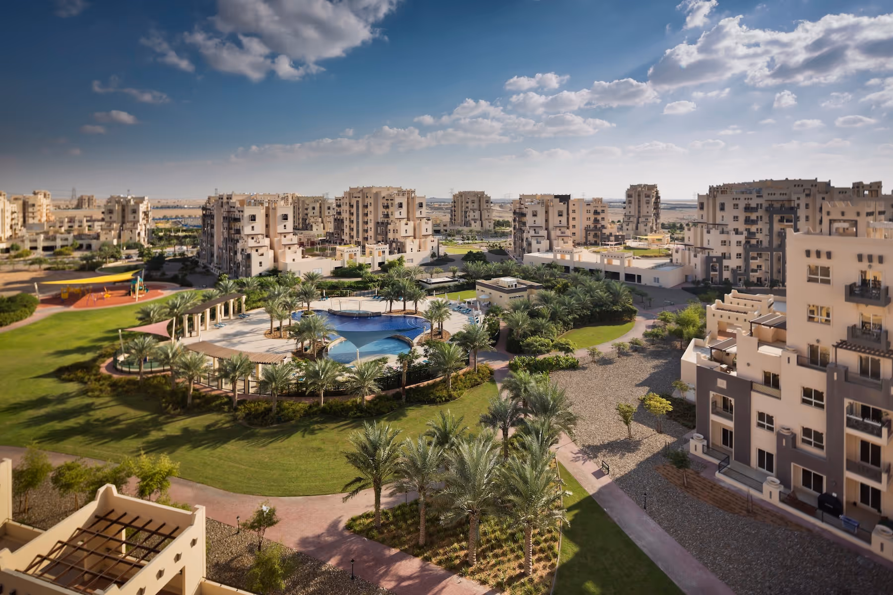 Aerial view of a residential community with mid-rise beige buildings, landscaped gardens, palm trees, and a central swimming pool area under a partly cloudy sky.