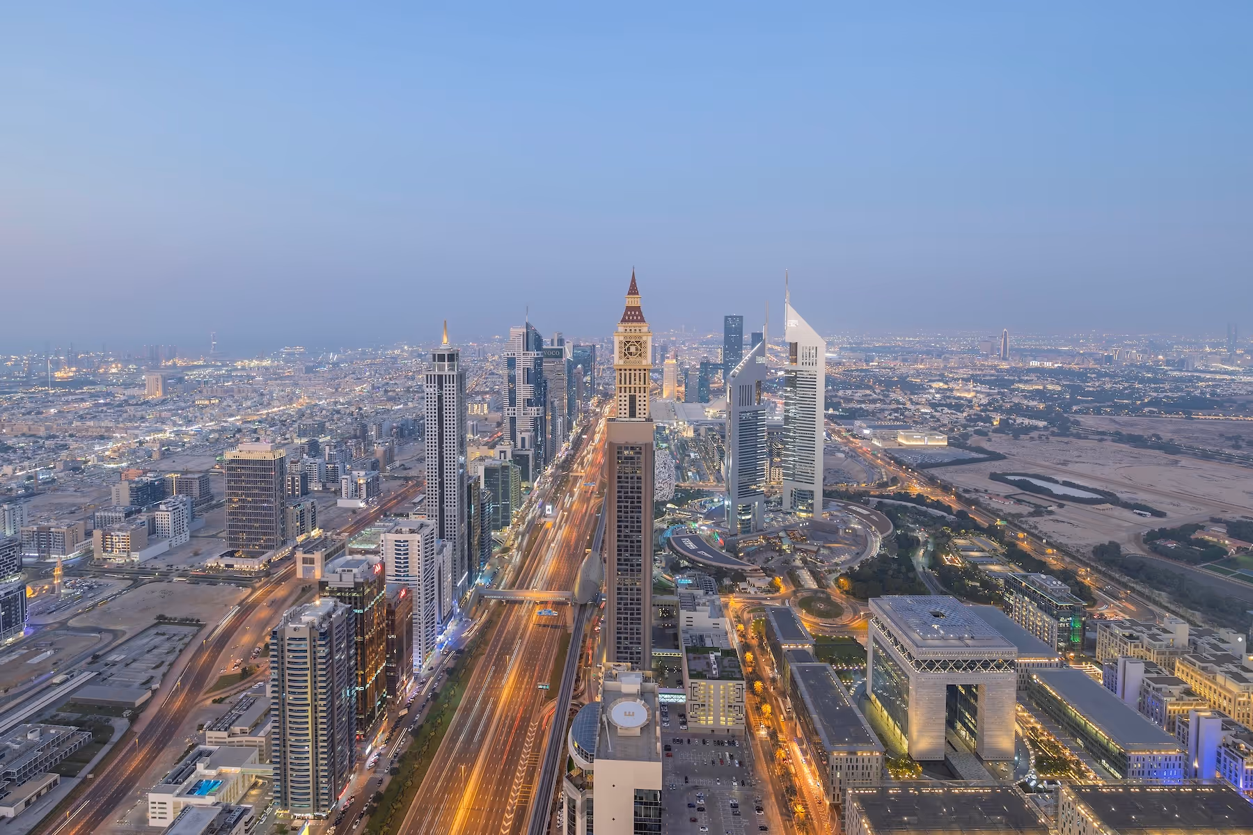 Aerial view of Sheikh Zayed Road in Dubai at dusk, featuring high-rise buildings including Emirates Towers and the clock tower in the foreground.
