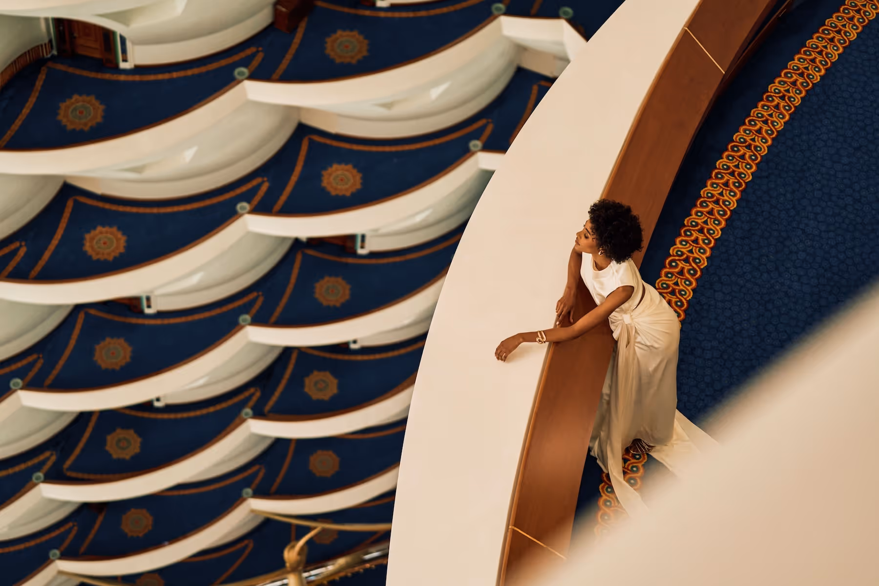 Woman in elegant white dress leaning on curved balcony railing overlooking patterned blue carpet and architectural interior.