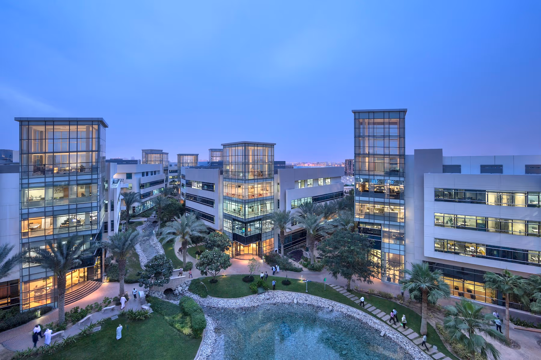 Modern office buildings surrounding a landscaped garden with palm trees and a small pond at dusk.