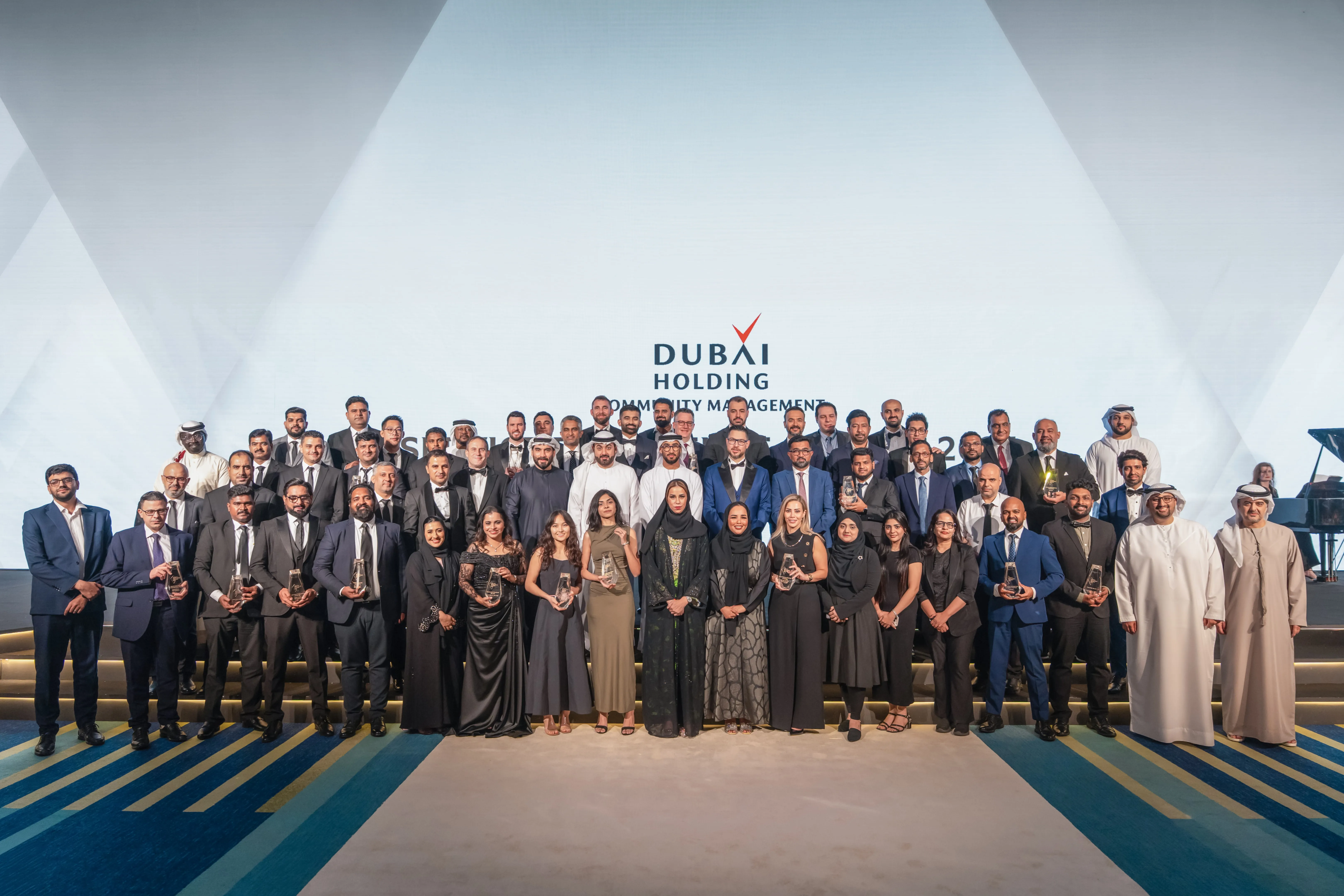 Group photo of formally dressed men and women, some holding awards, in front of a Dubai Holding Community Management backdrop at an event.