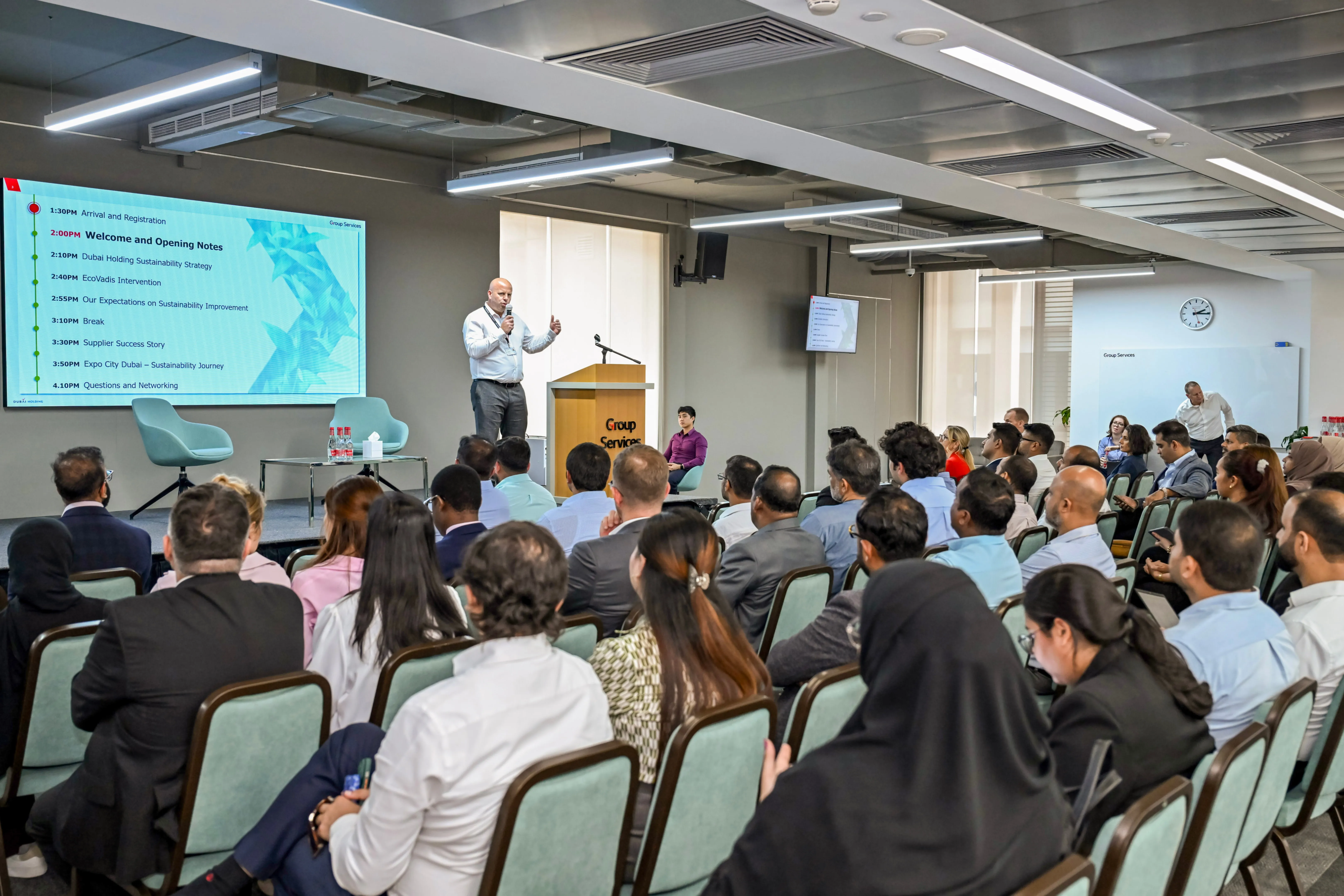 Speaker presenting sustainability strategy to a seated audience in a conference room with a schedule displayed on a large screen.