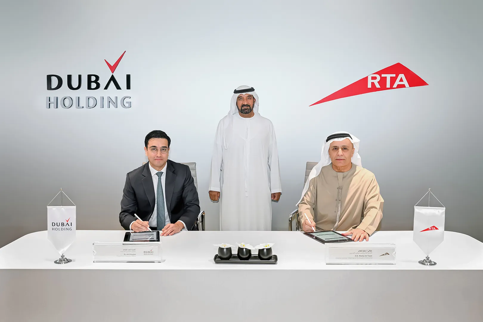 Two men sitting at a table signing documents with a man standing behind them, Dubai Holding and RTA logos on the wall.
