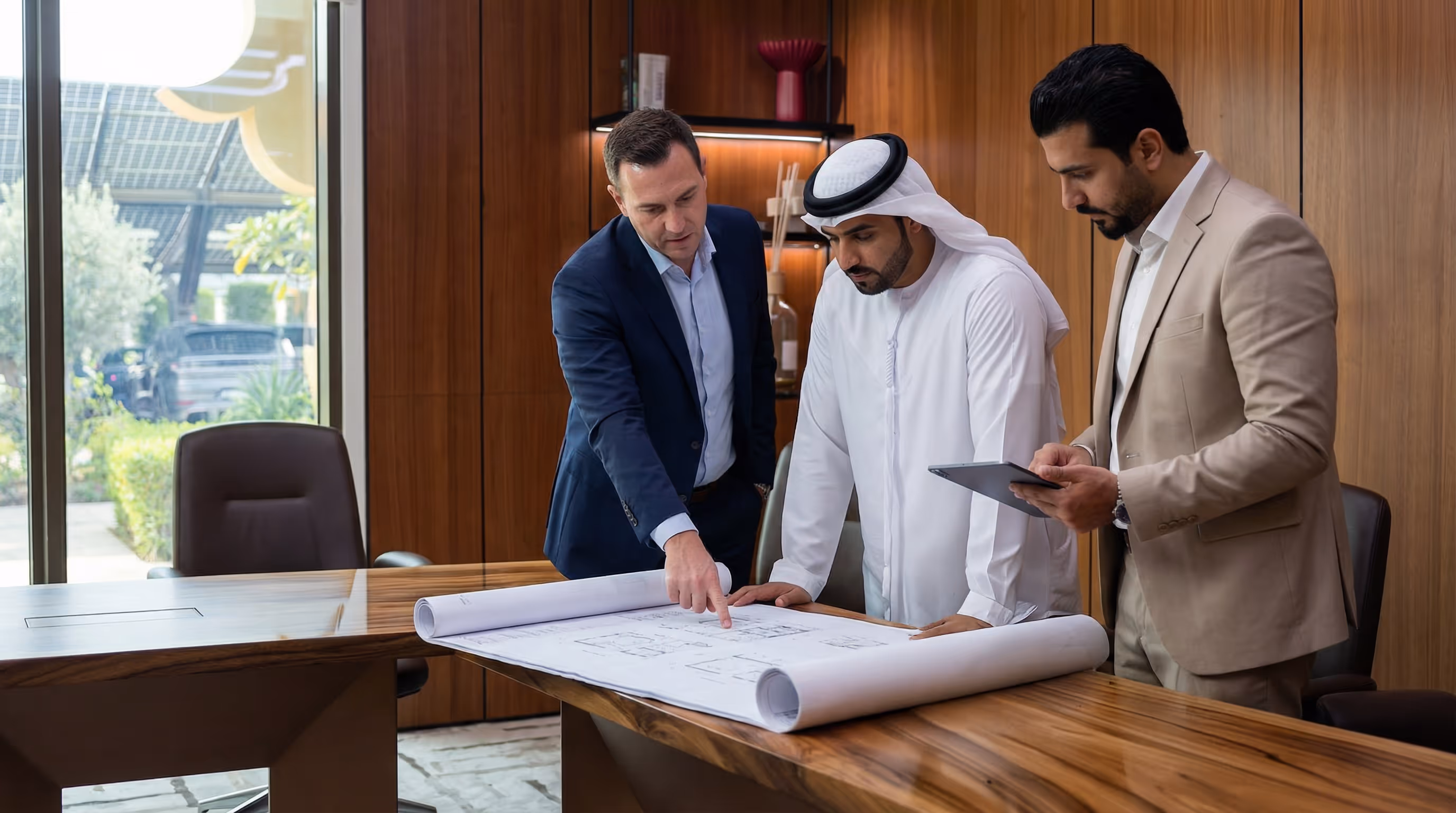 Three men in business attire reviewing architectural blueprints on a wooden conference table in a modern office.