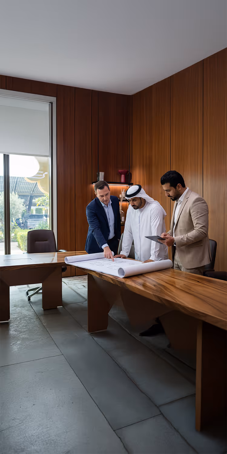 Three businessmen in an office discussing architectural blueprints on a wooden table; one points at the plans, another wears traditional Middle Eastern attire, the third checks a tablet.