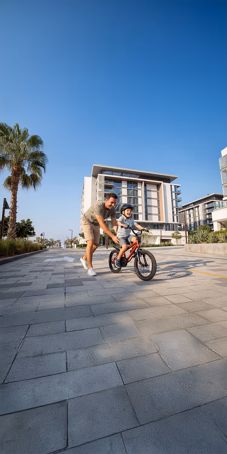 Father helping young boy ride a red bicycle on a paved street in a modern residential area with palm trees.