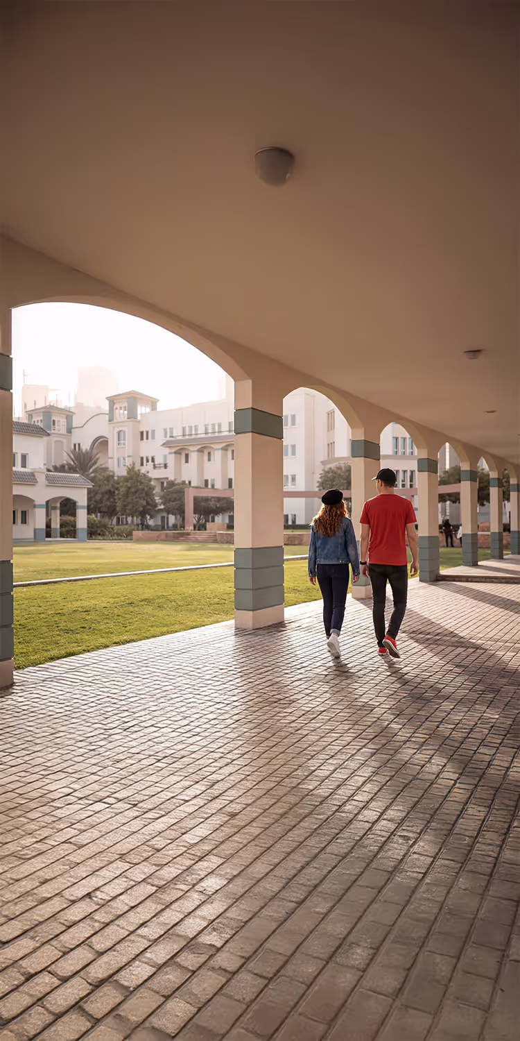 Two people walking under covered archway with green lawn and white buildings in the background.