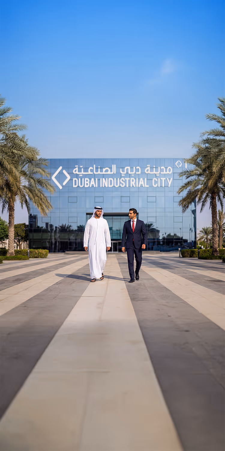 Two men walking on a wide pathway lined with palm trees in front of a glass building labeled Dubai Industrial City.