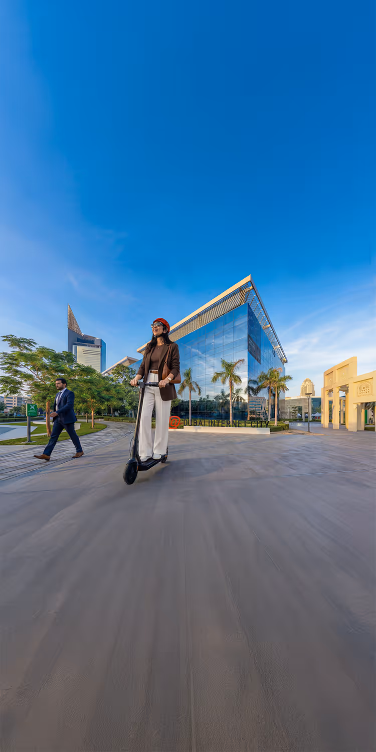 Woman wearing a red helmet riding an electric scooter near Dubai Internet City with modern buildings and palm trees in the background under a clear blue sky.
