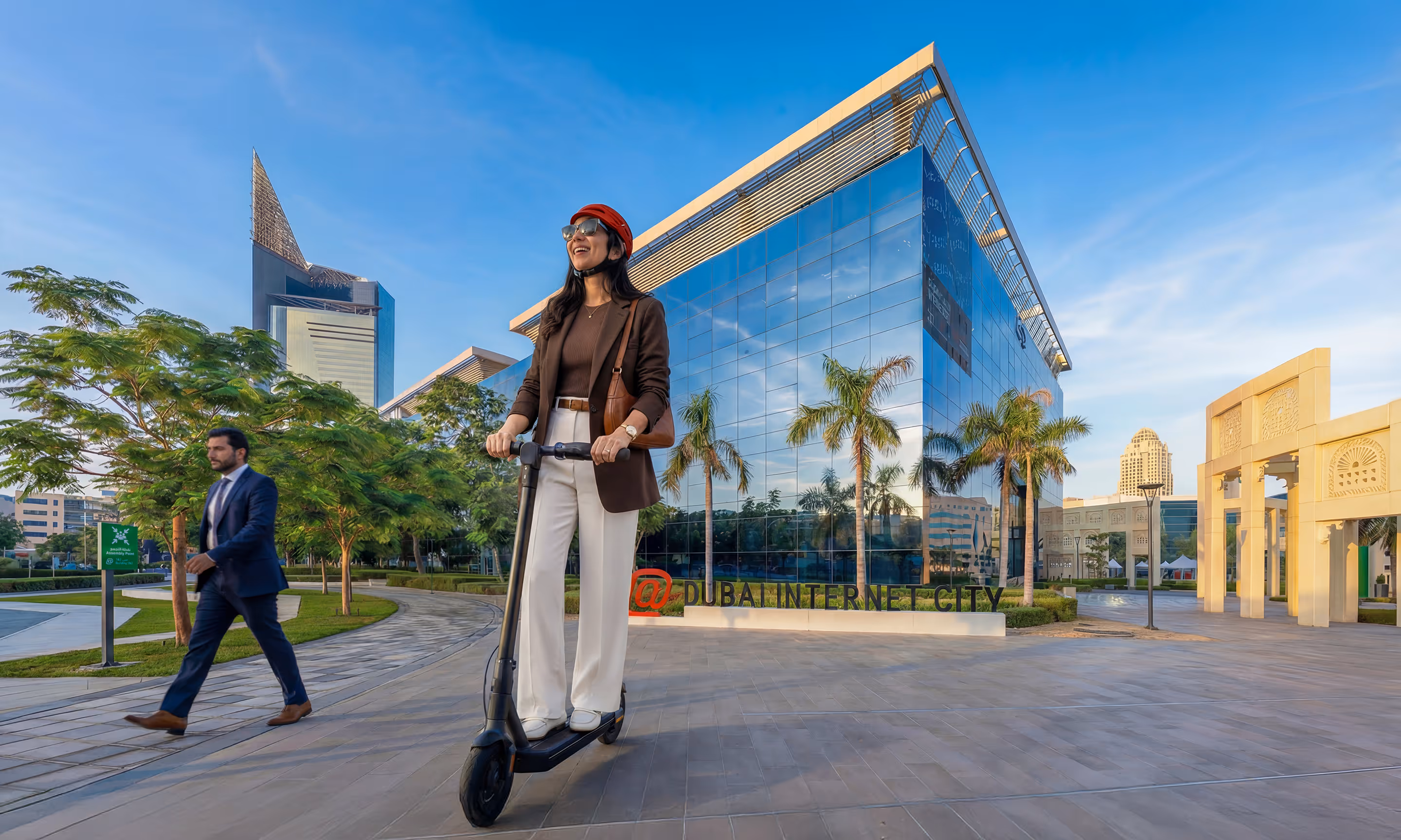Woman wearing a red helmet riding an electric scooter near a modern glass building with Dubai Internet City sign in the background during daytime.