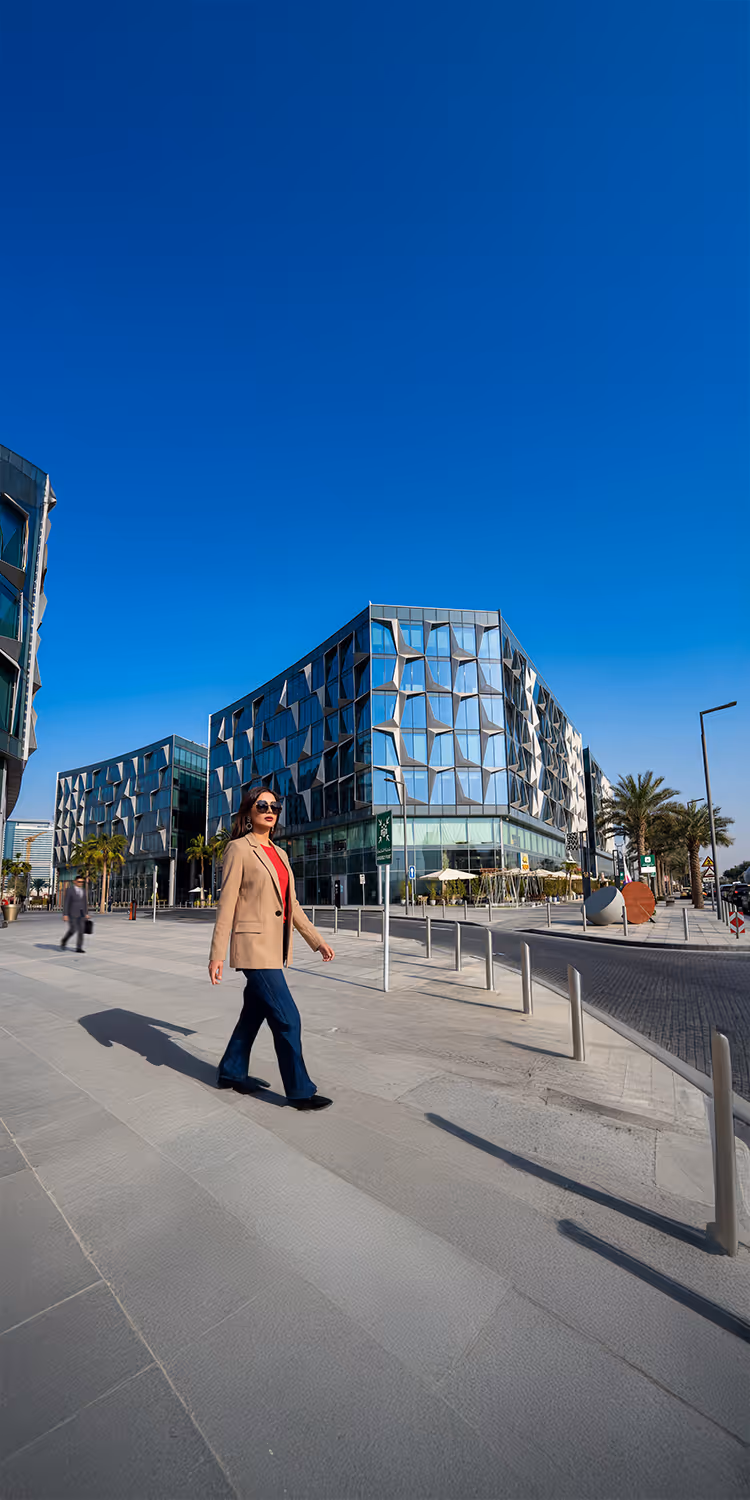 Woman in beige blazer and sunglasses walking on a modern city street with glass and metal buildings under a clear blue sky.