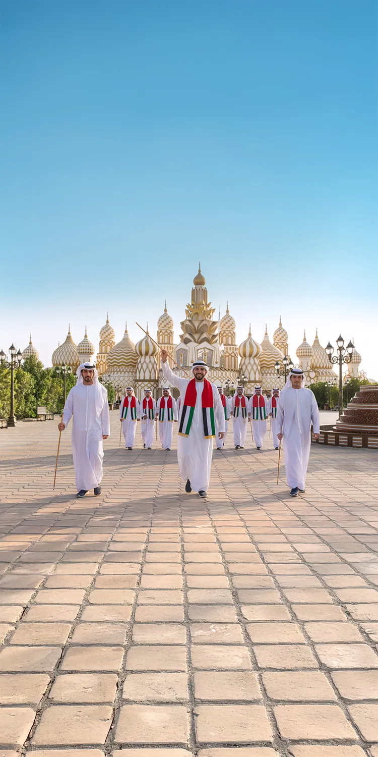 Group of men in white traditional Emirati clothing and scarves with UAE flag colors walking in front of the ornate, gold-topped Mosque of the Golden Domes under a clear blue sky.