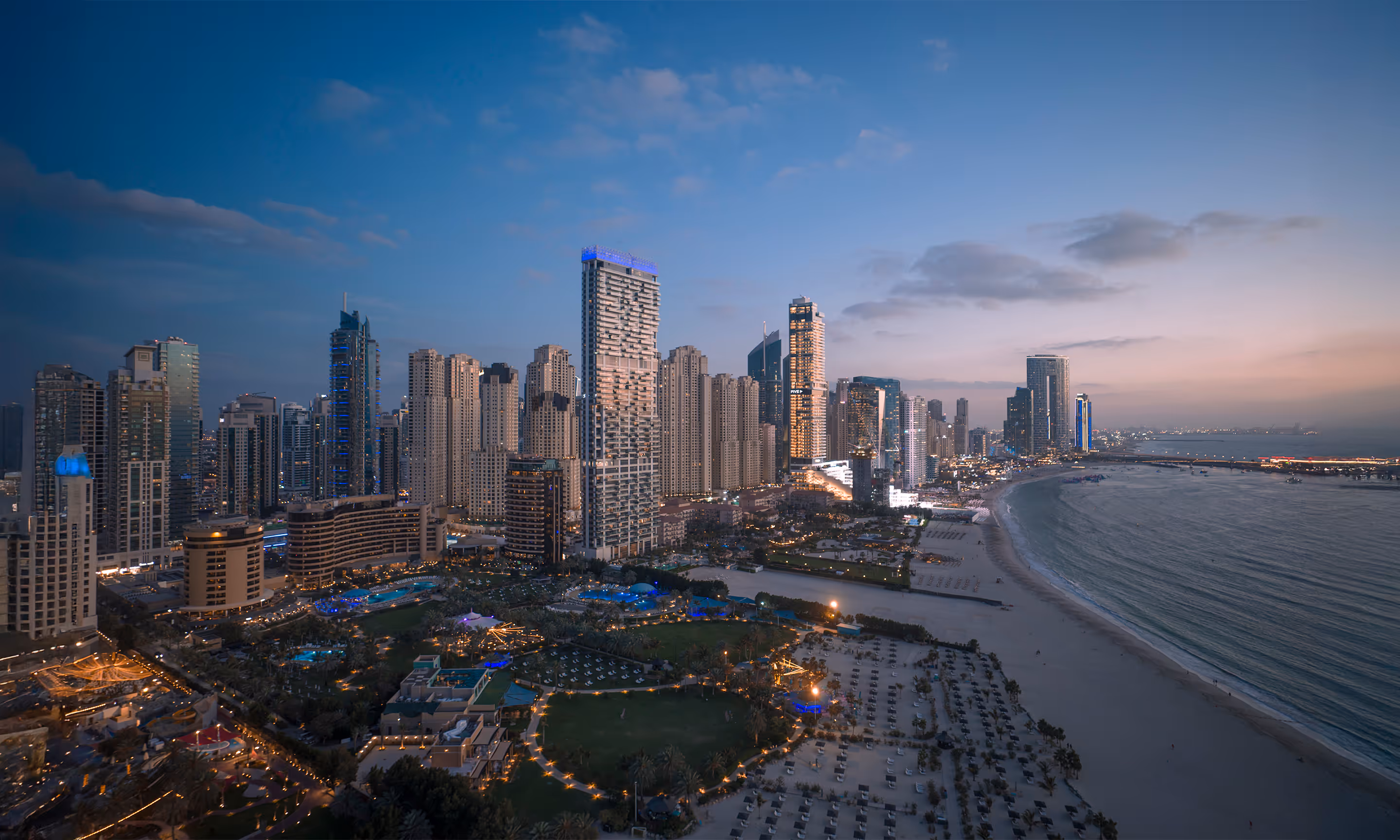 Evening cityscape of a modern coastal city with illuminated skyscrapers and a beach along the shoreline.