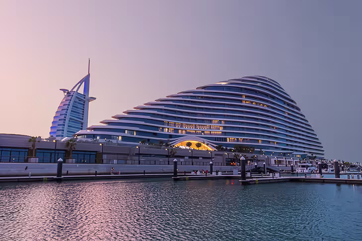 Modern hotel buildings by the waterfront at dusk with calm water reflecting the structures.