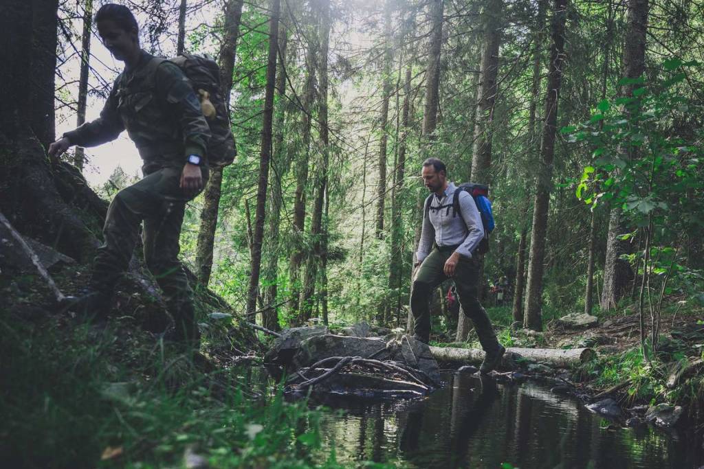 To turgåere med ryggsekker går på en skogssti ved en bekk omgitt av høye trær.