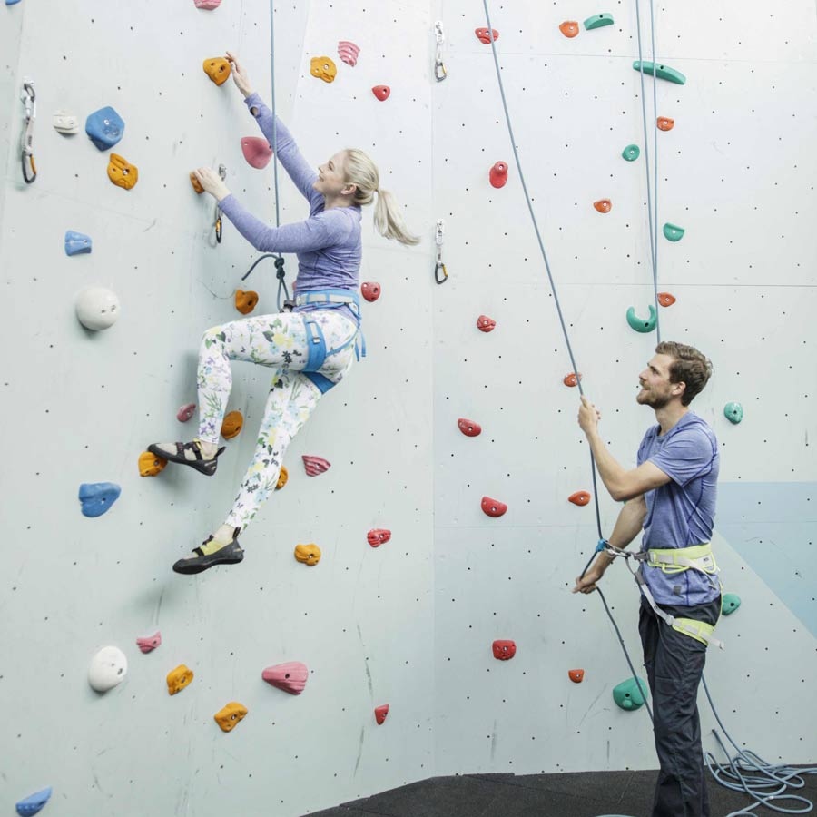 En kvinne klatrer på en innendørs klatrevegg mens en mann sikrer henne. Begge har på seg sikkerhetssele.