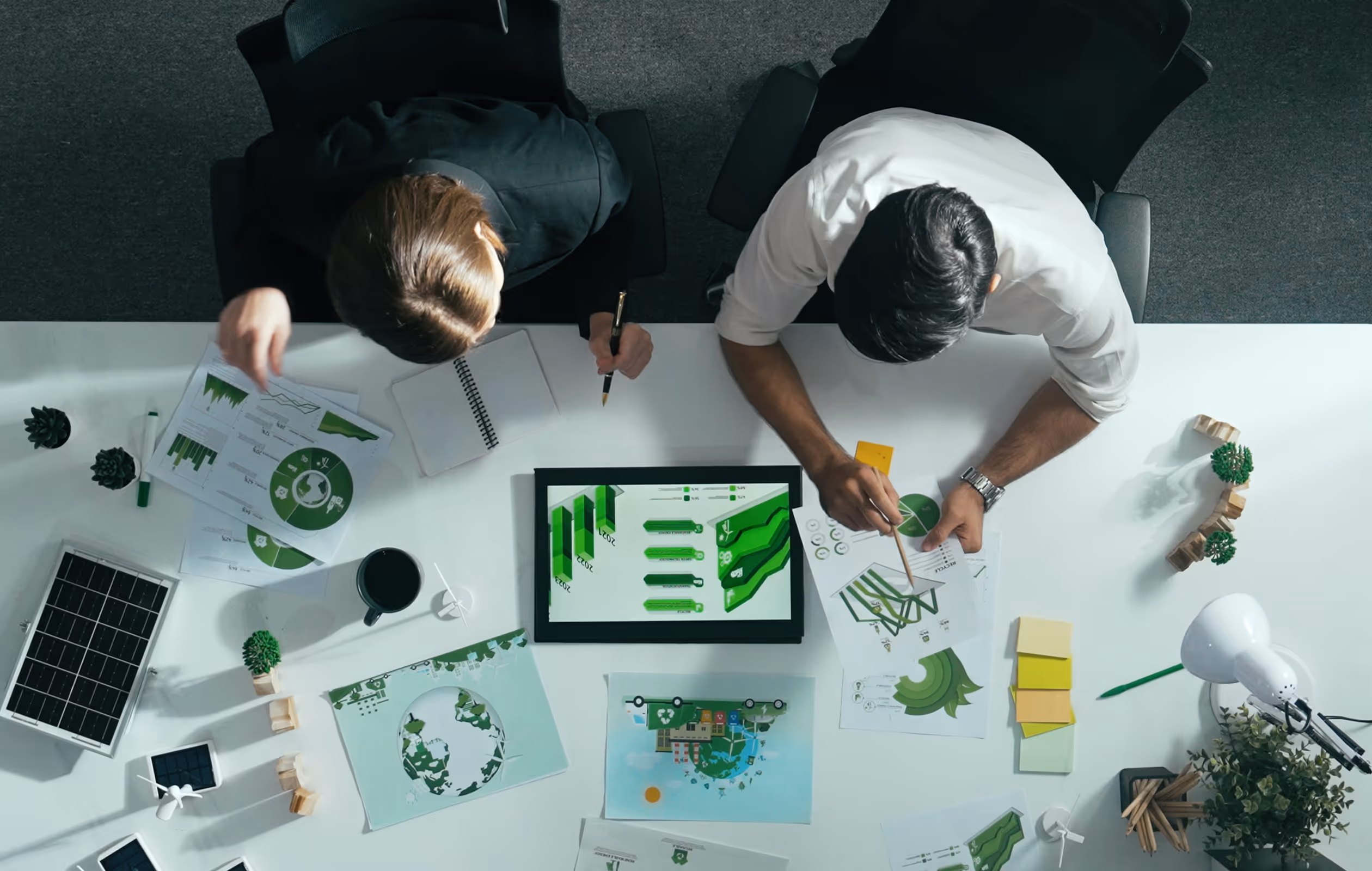 Top-down view of two people analyzing green-themed charts and graphs related to sustainability on a white desk.