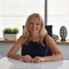 Smiling woman with blonde hair sitting at a white desk with a plant and sculpture in the background.