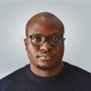 Man with glasses and a dark sweater looking at the camera against a plain light background.