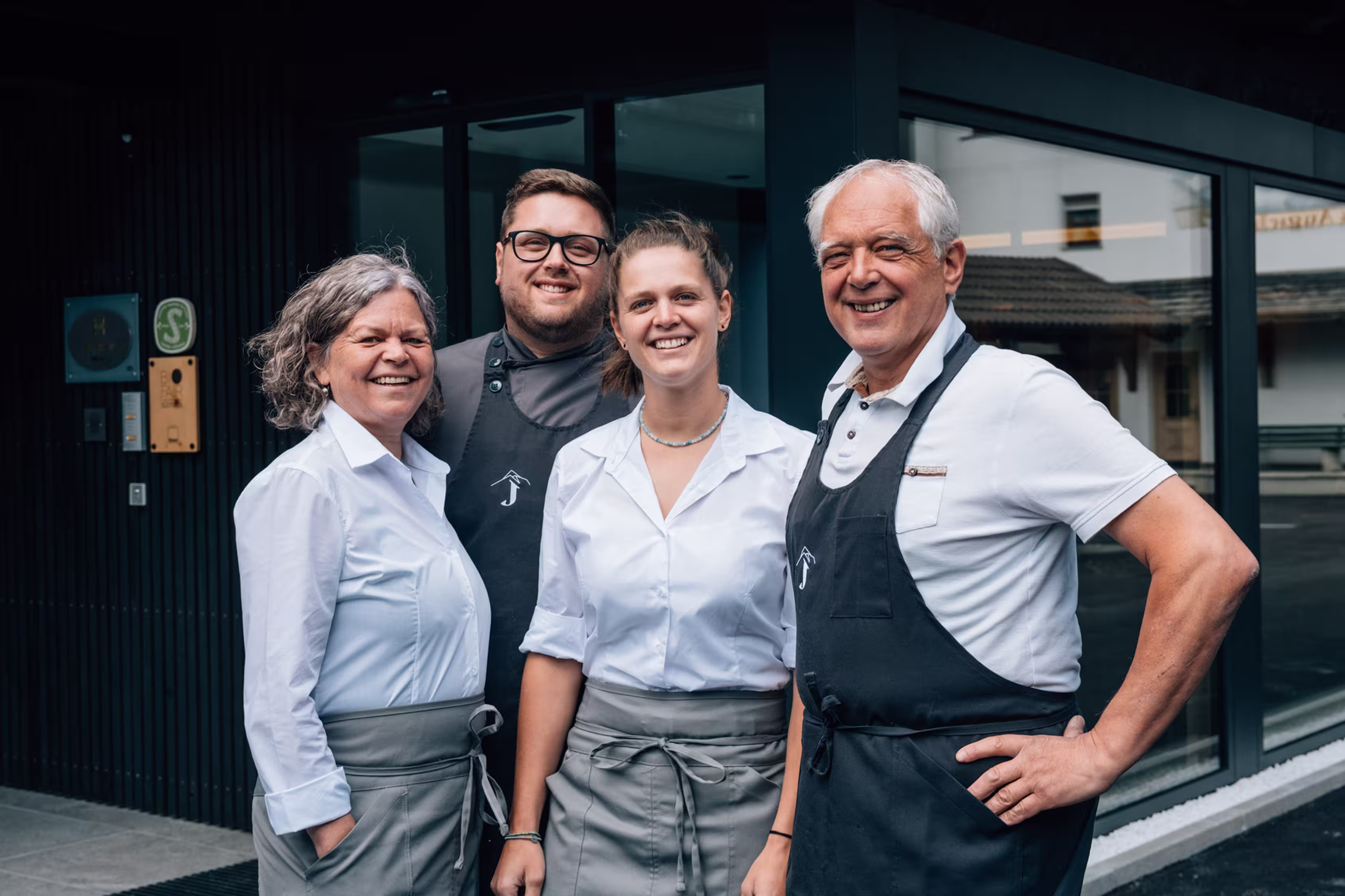 Four smiling restaurant staff wearing white shirts and aprons standing outside a building.