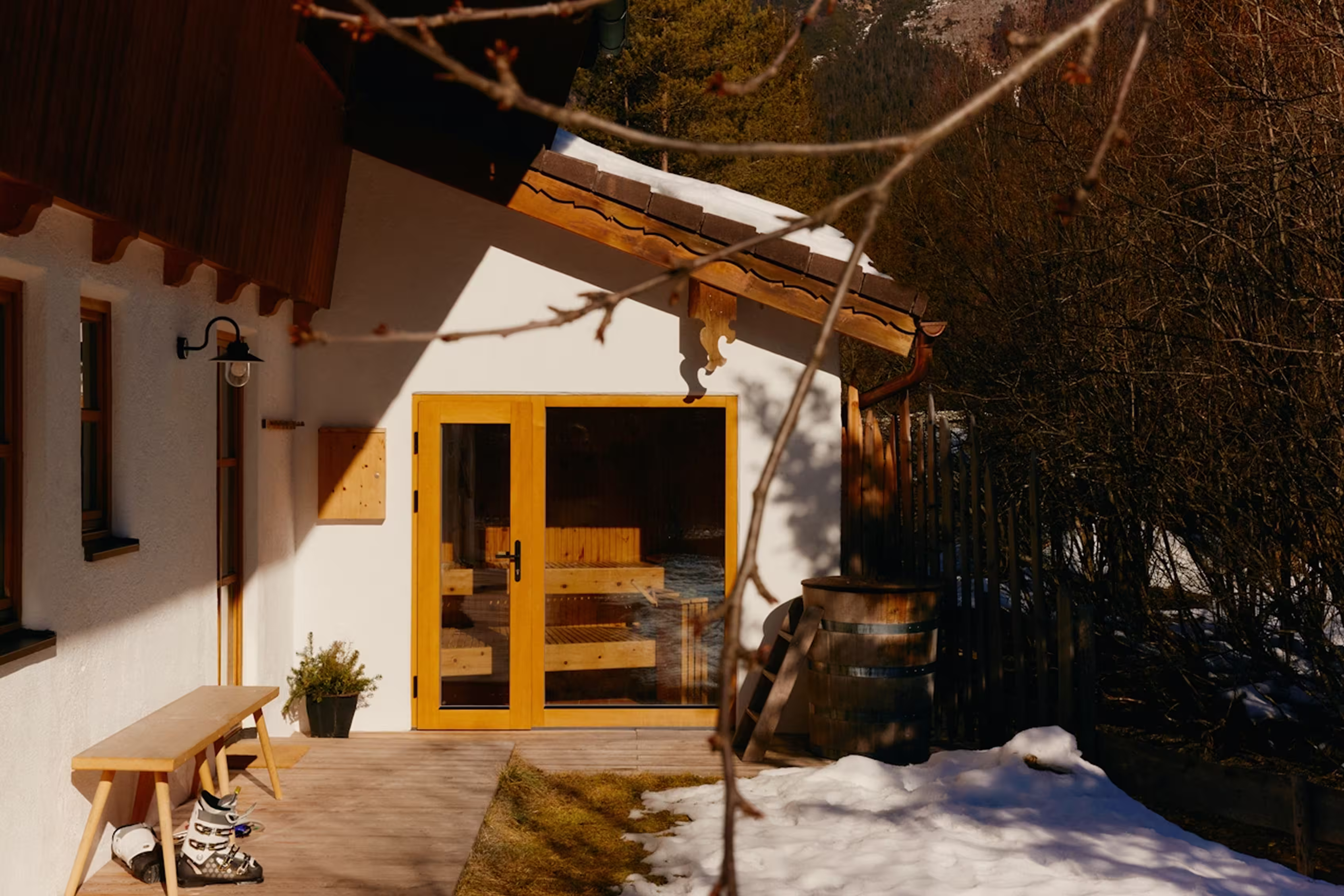 Wooden bench with ski boots outside a white house with yellow double glass doors and snow on the ground.