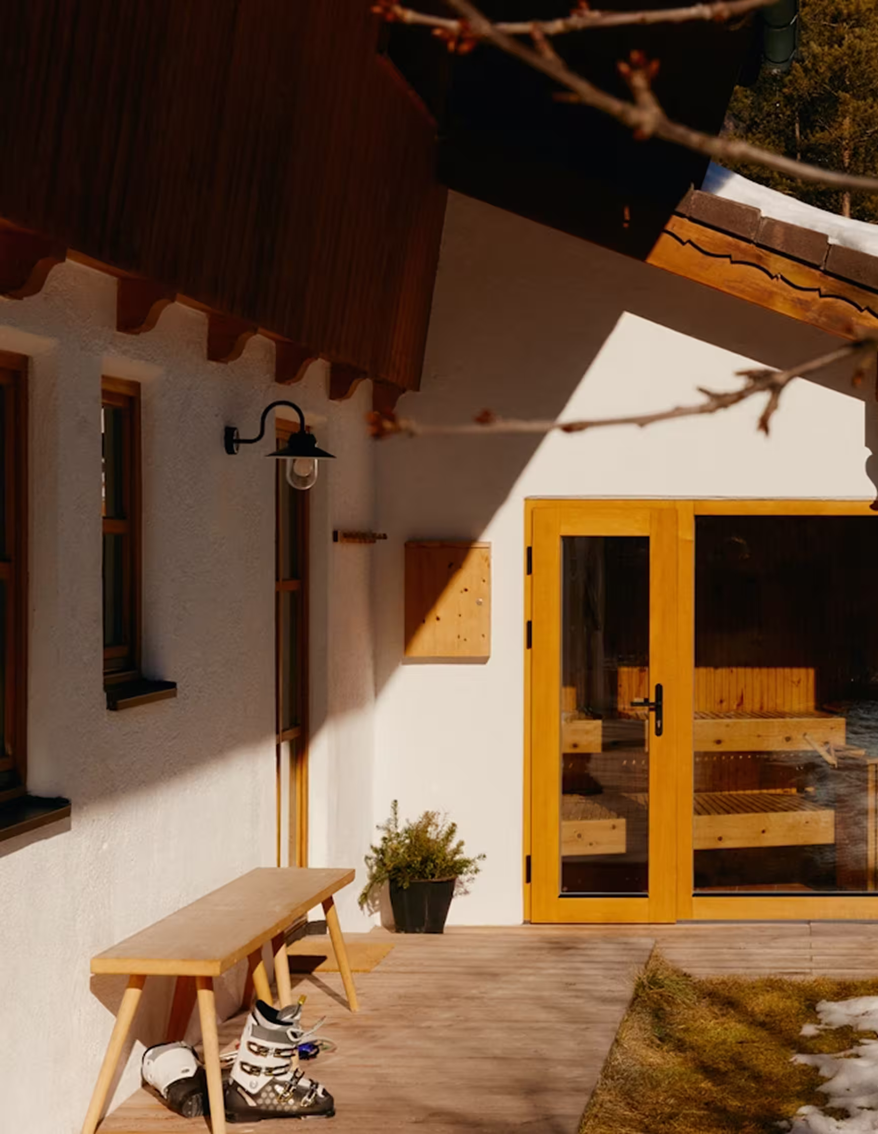 Wooden bench with ski boots underneath on a porch of a house with glass doors and some snow on the grass nearby.