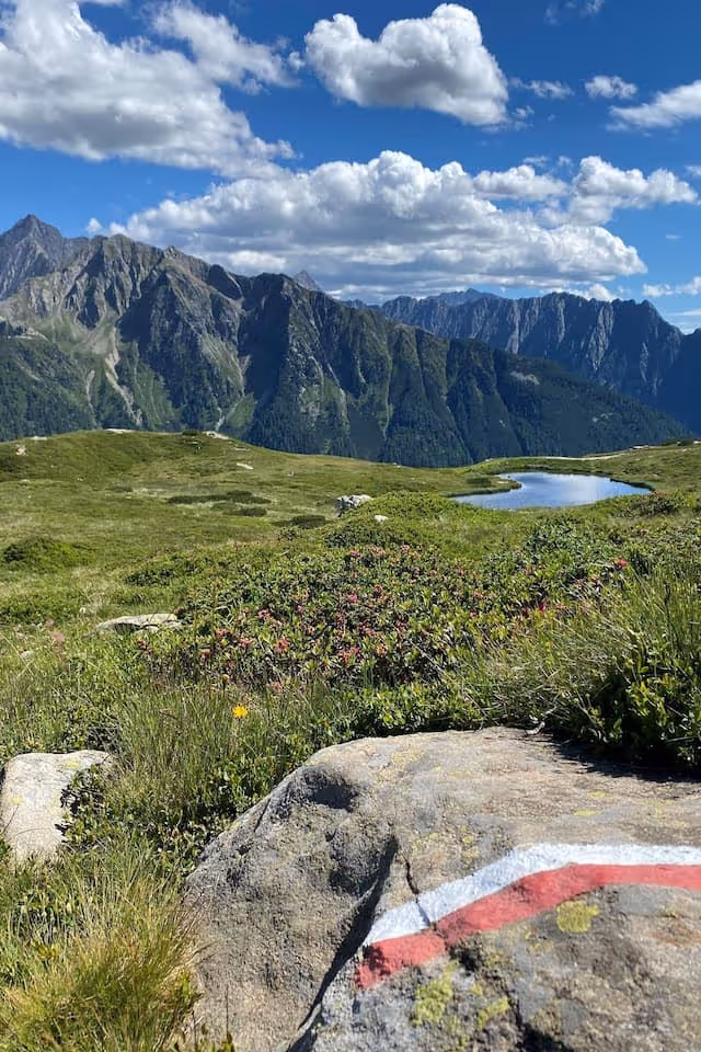 Berglandschaft mit grünen Wiesen, einem kleinen See, Felsen mit roten und weißen Markierungen und bewölktem blauem Himmel.