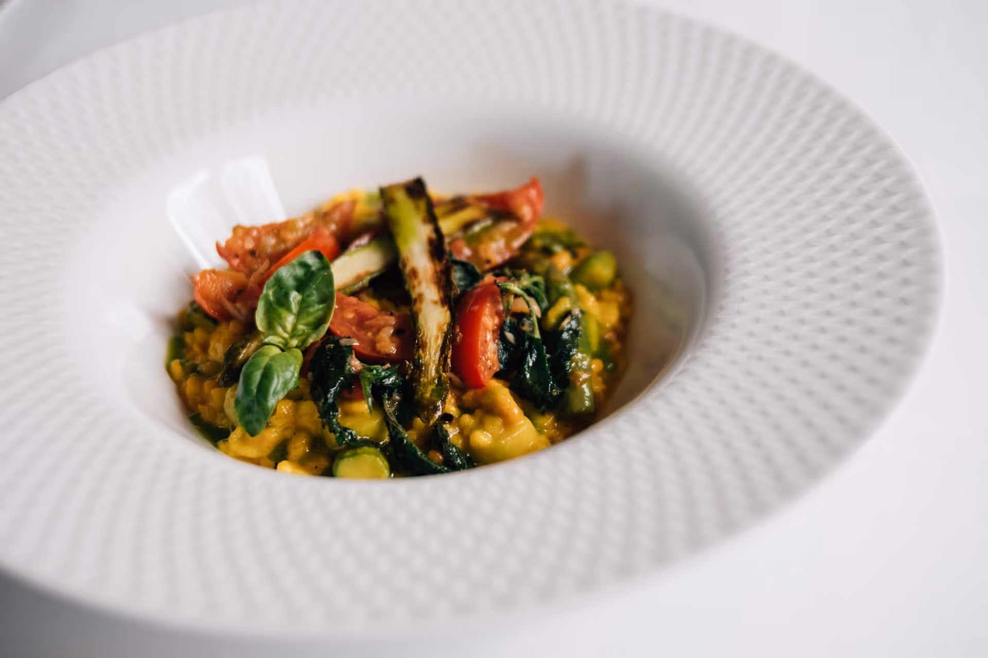Gelbes Risotto mit gegrilltem Spargel, Tomaten und frischen Basilikumblättern in weißem, strukturiertem Teller.
