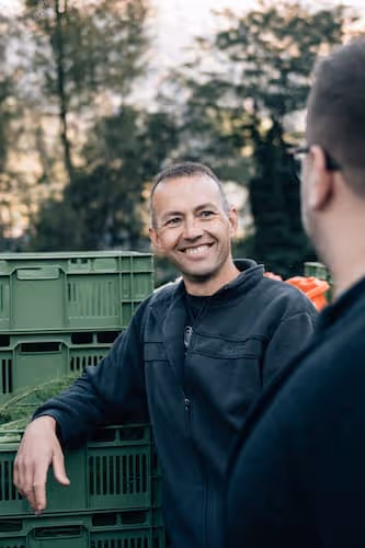 Zwei Männer unterhalten sich neben grünen Lagerkisten im Freien mit Bäumen im Hintergrund.