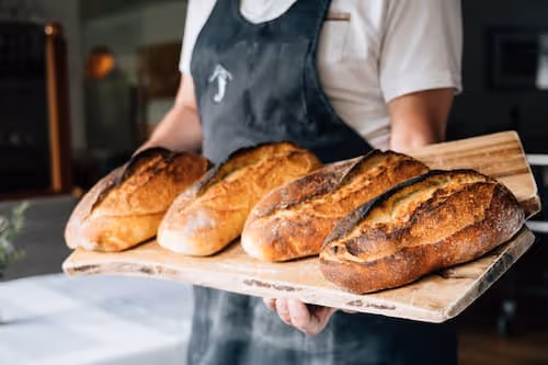 Person hält ein hölzernes Blech mit vier frisch gebackenen Broten.
