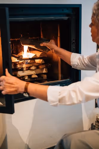 Person fügt ein Stück Holz in einen Kamin mit brennendem Feuer ein.