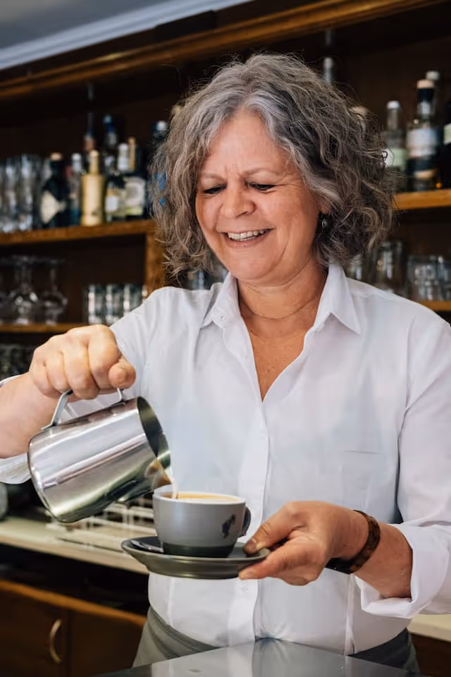 Ältere Frau mit grauen Haaren gießend Kaffee aus einer Metallkanne in eine graue Tasse in einem Café.